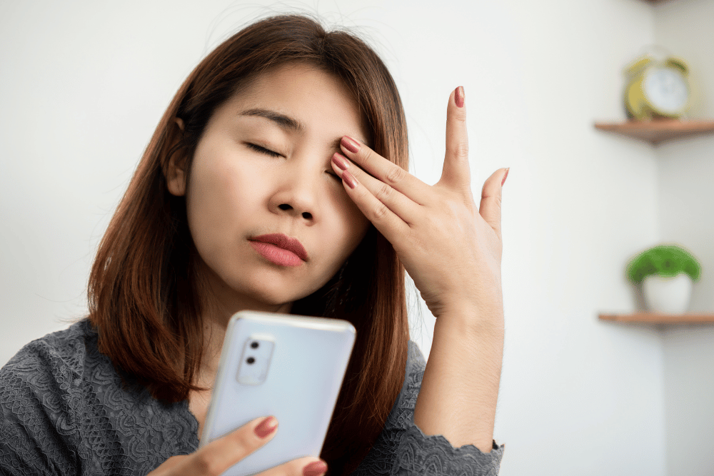 a woman holding a hand to her eye in discomfort while holding her phone